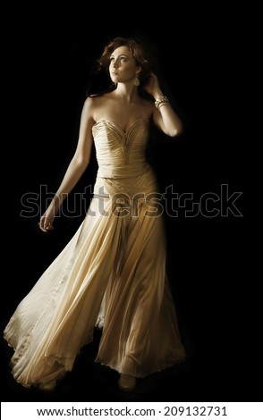 Gorgeous brunette woman wearing a flowing couture strapless evening dress wearing gold shoes and bronze makeup, posing with her hand in her auburn hair