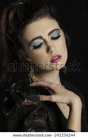 Lovely eighties brunette woman in colorful graphic makeup posing with her hand on her shoulder while looking downwards.