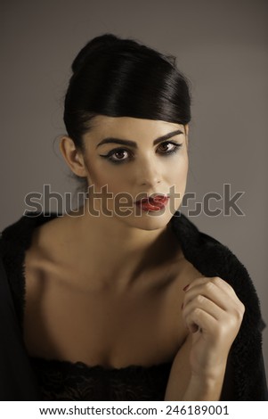 Portrait of dark haired woman wearing black cape, dark eyeliner and makeup, with red lips, looking seductively at viewer with her sultry brown eyes