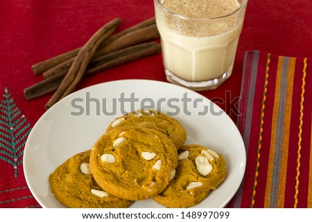 A plate full of soft, chewy pumpkin spice cookies is served with a glass of traditional holiday egg nog topped with nutmeg on a Christmas themed place mat.
