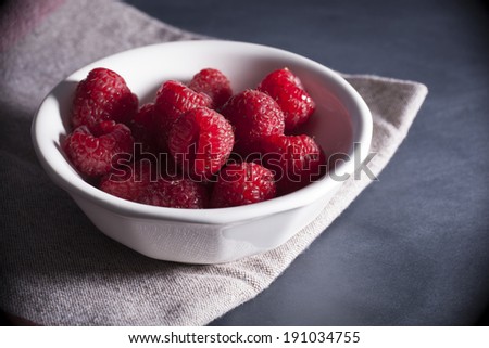 A white bowl of bright red fresh raspberries sits atop a rustic looking napkin and black surface