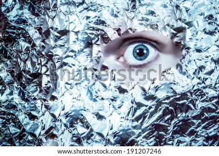 An eye peeks out of  a mask of tin foil.  Looks frightening and surreal.  Split colortone