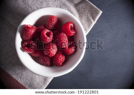 A white bowl of bright red fresh raspberries sits atop a rustic looking napkin and black surface