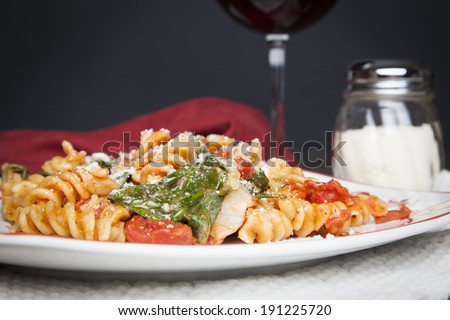 A red and white plate filled with Fusilli pasta, prepared with fresh baby spinach, diced tomatoes and parmesan cheese; a glass of red wine and shaker of parmesan cheese sit off to the side