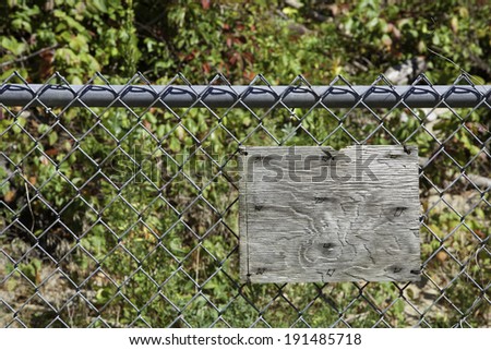 An old blank wooden sign hangs on a gray chain link fence; wooded area and brush behind fence