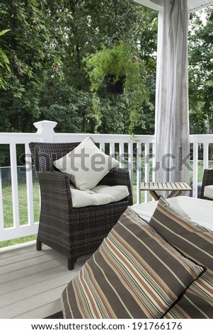 Two resin wicker chairs sit on a covered deck; they have cream colored cushions and pillows;  canvas curtains hang in the background; a white wicker chest serves as a coffee table