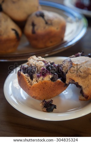 A whole blueberry muffin, torn apart, sits on a white plate on a dark wood tabletop.  A tray of muffins along sits in the background.