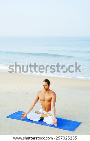 Long hair athletic man with no shirt meditatting on blue mat at the beach Padmasana - Lotus position