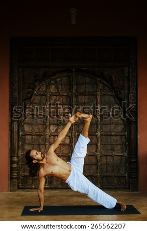 Indian ethnicity young man wit a strong body showing yoga pose with leg up holding big toe (asana) in front of old oriental style door.
