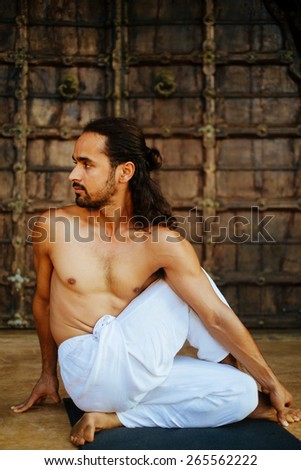 Indian ethnicity young man wit a strong body showing half seated spinal twist yoga pose (Ardha-Matsyendrasana) in front of old oriental style door.