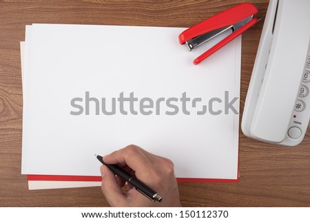 A hand writing on sheets of paper, a white phone and a red stapler on wooden texture imitating a office desk.