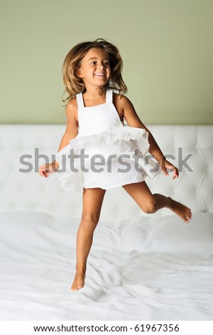 A happy cute young girl in white dress having fun jumping on bed
