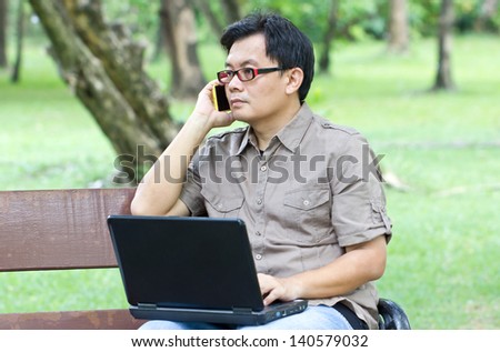 Asian man with mobile phone working with laptop in the park.