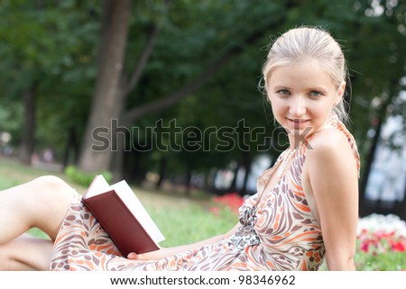 young woman reads the book in hands sits on a green grass in park in a light dress and smiles