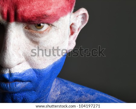 Man with his face painted with the flag of Netherlands.  The man is serious and photographic composition leaves only half of the face.