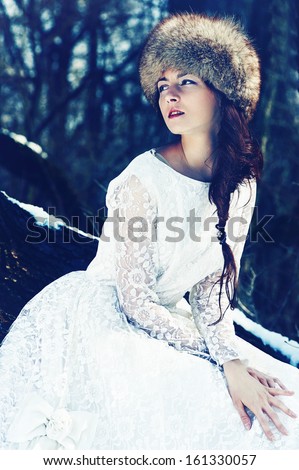 Fashion young beautiful brunette in fur hat and white luxury dress sitting outdoor in winter