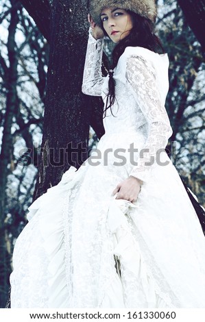 Fashion portrait of young serious brunette woman in fur hat and white luxury dress in winter wood background