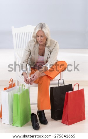 senior woman sitting on a chair surrounded by shopping bags and rubbing her sore feet.