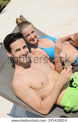 happy young couple cheering with champagne in deck chairs by hotel resort pool