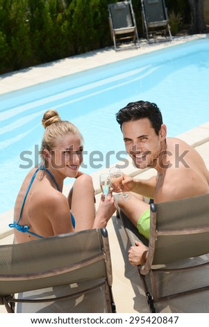 happy young couple cheering with champagne in deck chairs by hotel resort pool