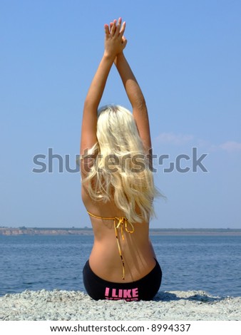 The girl-blonde sits on coast of a gulf, having lifted hands upwards