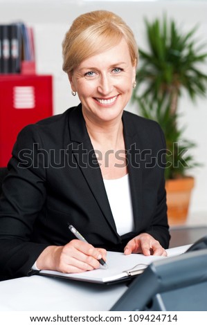 Seated female secretary writing on notepad in office and looking at you