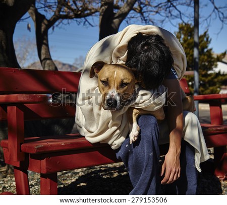 Man with dog wrapped in blanket sitting on red bench under tree slouched over dog