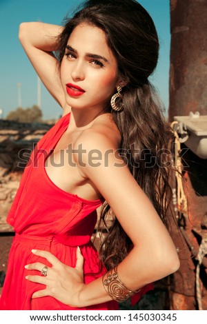 red passion. adorable brunette woman in red dress with long brown hair and golden accessories posing on a sunny day near a bridge . outdoors shot. soft sunny colors