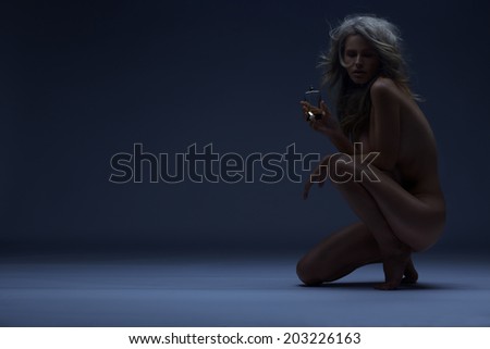 Art photo of naked blonde with fragrance.  Naked woman with glossy skin holding perfume. Long shiny blond hair. Cosmetics concept.Toned in warm colors. Shot in studio, horizontal.