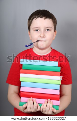 Boy with a pen in his mouth holding a stack of books, gray background.