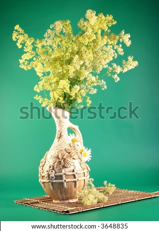 On a wooden napkin a jug with bouquets of yellow field colors cost. It is photographed on a green background.