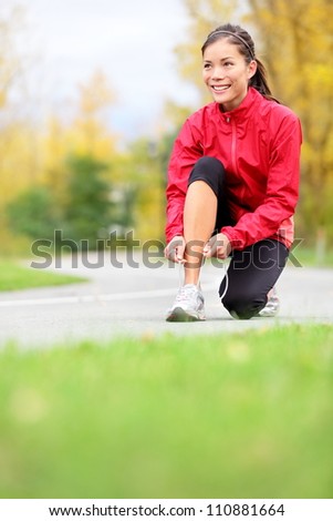 Runner woman tying running shoes outside in fall. Beautiful young fitness model smiling happy in casual jogging clothing.