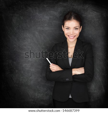 Woman teacher or business woman at blackboard holding chalk standing in suit by blackboard teaching or giving lecture. Young female professional portrait. Mixed race Asian Caucasian female model.