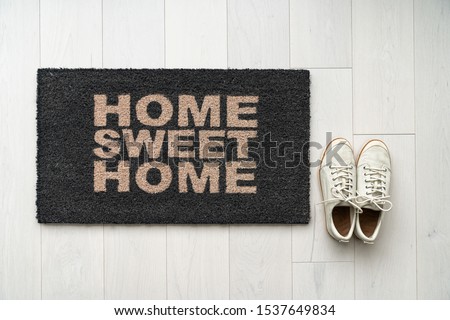 New home moving in door mat entrance welcome doormat with text writing HOME SWEET HOME and white sneakers of happy condo homeowner. Top view of rug and wooden floor.