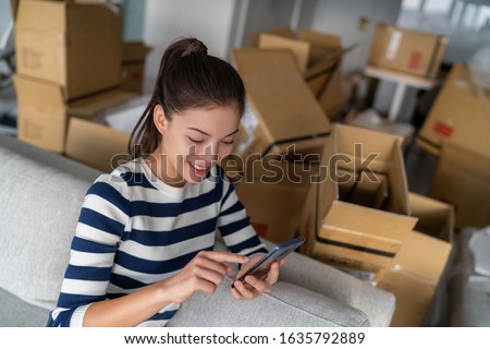 Homeowner Asian woman using cellphone mobile phone for indoor mess with many open packages moving boxes for move in new apartment. Happy girl in condo house.