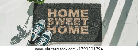 Home sweet home door mat at house entrance with women's sneakers of woman that has just arrived. New condo homeowner moving in with plant. Panoramic crop.