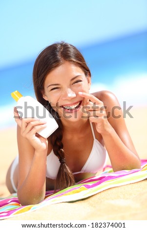 Suntan lotion woman applying sunscreen solar cream laughing having fun. Beautiful happy cute woman asian applying suntan cream from a plastic container to her nose with ocean in background.