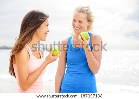 Healthy lifestyle women eating apple after running. Two beautiful girlfriends talking enjoying fruit after jogging training on beach. Caucasian woman and Asian woman in their 20s smiling happy.