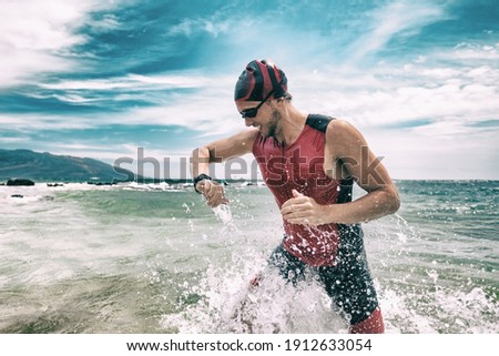 Triathlete swimmer looking at sport watch app using smartwatch during triathlon. Swimming man running out of ocean swim checking heart rate on smart watch. Professional athlete training for ironman.