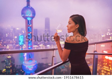 Luxury Shanghai lifestyle Asian woman drinking white wine in on China Travel. Elegant model in black dress at terrace view of the city, high end living.