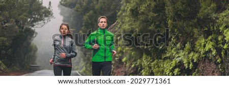 Runners jogging outside on wet trail running in the rain in autumn fall outdoor cold season. Panoramic of couple athletes training together.