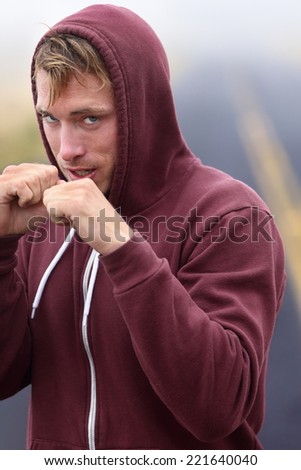Fitness boxer boxing outside on road in hoodie. Man punching and hitting looking at camera. Fit fitness boxer sweating in sweatshirt outside. Caucasian male fitness model in his 20s.