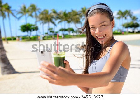 Fitness selfie woman drinking green vegetable smoothie taking self portrait photograph with smart phone after running exercise workout on beach. Healthy lifestyle with fit Asian Caucasian girl.