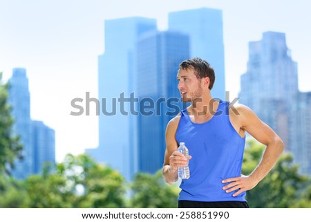 Sport man drinking water bottle in New York City. Male runner sweaty and thirsty after run in Central Park, NYC, Manhattan, with urban buildings skyline in the background.