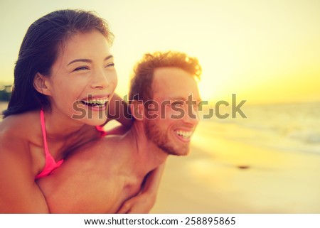 Happy beach fun multicultural couple - summer love in sunset. Young adults piggybacking playful and laughing on hawaiian beach, Asian woman and Caucasian man. Multiethnic mixed race relationship.