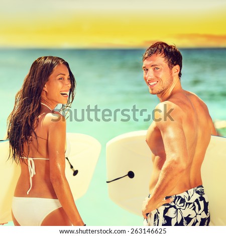 Surfers on beach having fun in summer. Surfer woman and man with boogieboard smiling happy on beach on Hawaii. Multiracial couple Asian woman and Caucasian man in outdoor water activity during travel.