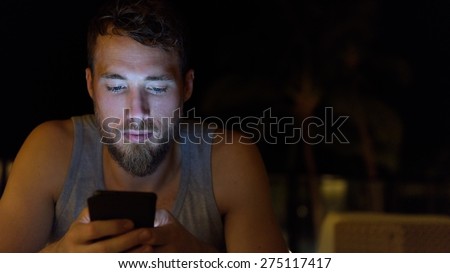 Man using smartphone at night browsing internet updating social media. Young male with beard using mobile cell smart phone outdoors in the dark in summer.