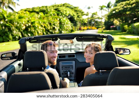 Happy couple in car on summer road trip travel. Multiracial young couple carefree on holidays driving a convertible cabriolet automobile on the roadway in the city looking back at camera.
