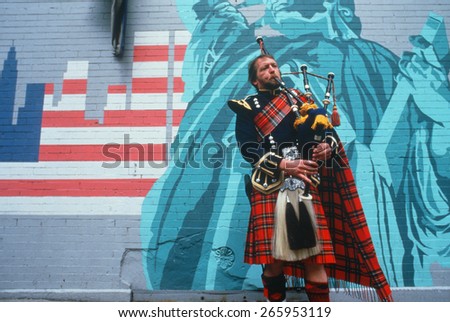 A man in kilt playing bagpipes in front of a patriotic mural, St. Patrick\'s Day Parade, NY City