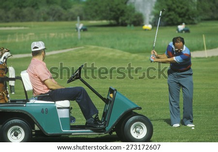 Golfer swinging club while man in cart looks on